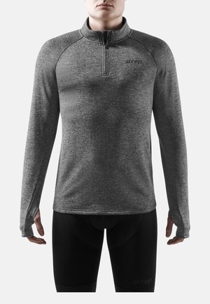 Mann trägt eng anliegendes graues langärmliges Sportoberteil mit Daumenlöchern und schwarze Kompressionsshorts, steht vor einfarbigem Hintergrund.