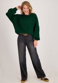 Vrouw glimlachend, draagt een groene oversized trui, vervaagde zwarte wijde jeans, een bruine riem en bruine sneakers met witte strepen.