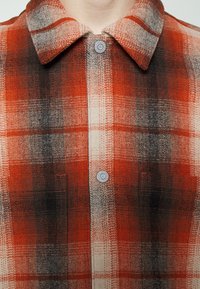 Rood en grijs geruite shirt met een knoopsluiting, drukknoopjes en twee voorzakken van een gladde, gestructureerde stof.