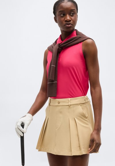 Kvinde iført en lyserød ærmeløs polo med en brun sweater lagt over skuldrene, beige plisseret nederdel, hvid golfhandske, der holder en golfkølle.