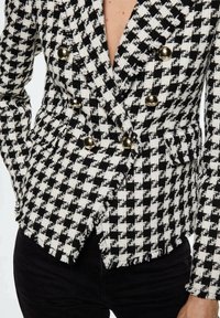 Sako s černobílým vzorem houndstooth vyrobené z texturované látky, vybavené zlatými knoflíky a přiléhavým siluetou s třásněmi.