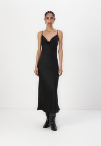 Svart satin midi-kjole med cowl-halsringning og tynne stropper, som har en glatt tekstur og en litt utsvingt nederkant. Brukt med svarte høye støvler.