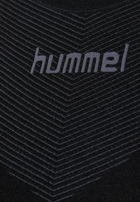 Prikup črne tkanine z tankimi svetlo sivimi chevron črtami in natisnjenim imenom blagovne znamke "hummel" v svetlo sivi barvi.