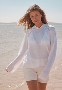 Sweat léger blanc à capuche avec manches longues et ourlet côtelé, associé à un short clair, sur fond de plage.