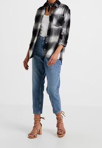 Camisa de cuadros en blanco y negro con botones, jeans de cintura alta en azul y sandalias de tiras en color beige con tacones cuadrados. Los accesorios incluyen una bufanda negra.