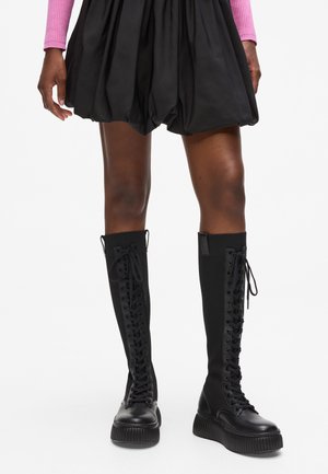 Mustad muste põlvekonnani saapad siledast nahast varbaga, ribastest kangast vars ja paks kummist platvorm. Esiküljel on paelad ja matt viimistlus.