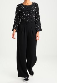 Blusa nera a maniche lunghe con pois bianchi, dotata di polsini a campana; abbinata a pantaloni neri a vita alta a gamba larga e sneakers neri.