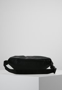 Černá kožená kabelka přes rameno obdélníkového tvaru, se zipovou kapsou, nastavitelným popruhem a hladkým povrchem. Má minimalistický design.