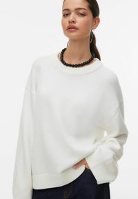 Pull blanc en maille avec un col rond et des épaules tombantes. Poignets et ourlet côtelés. Associé à un collier de perles noir.