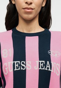 Πλεκτό πουλόβερ με οριζόντιες ρίγες σε ναυτικό μπλε και ροζ. Φέρει το λογότυπο "GUESS JEANS" και τον αριθμό "81" σε κρεμ χρώμα.