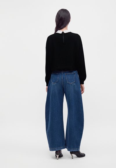 Personne aux longs cheveux foncés, vue de dos, portant un pull noir, un jean bleu à jambes larges et des bottines noires à talons.