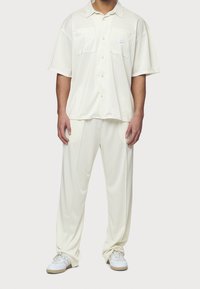 Kremowa koszula na guziki z dwoma przednimi kieszeniami, w zestawieniu z kremowymi spodniami. Oba elementy mają gładką fakturę i luźny krój. Do tego białe trampki.