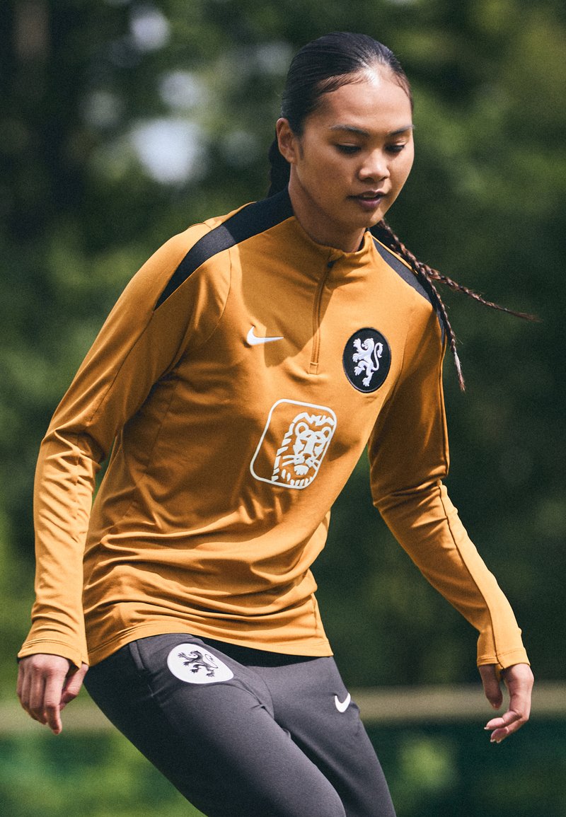 Long-sleeved amber training jersey with a black accent, featuring a lion logo and sponsor graphic, paired with dark gray athletic pants.