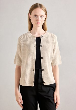 Femme portant un cardigan en tricot beige à manches courtes sur un haut noir et un pantalon noir, se tenant devant un fond gris clair uni.