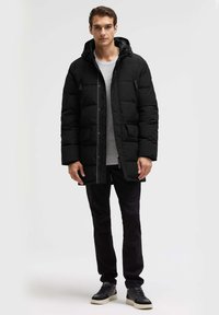 Schwarze Steppjacke mit Kapuze, vorne mit Reißverschluss, mehreren Taschen und gesteppter Textur. Kombiniert mit dunklen Hosen und schwarzen Sneakern.