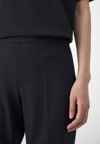 Black textured pants with a smooth waistband and subtle pleats, worn with a black t-shirt, focus on the waist and hand details.