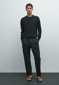 Black long-sleeve shirt with a round neckline, paired with black tailored pants and black flip-flops, standing against a light gray background.