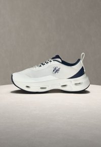 Baskets de sport blanches et bleu marine avec fermeture à lacets à l'avant, semelle épaisse et rembourrée avec des poches d'air, et logo brodé "EA" sur le côté.