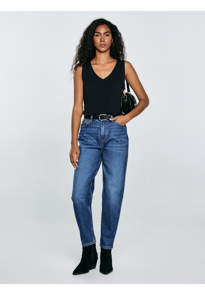 Vrouw met lang krullend haar, gekleed in een zwart mouwloos topje, blauwe high-waist jeans, zwarte laarzen en met een zwarte schoudertas.