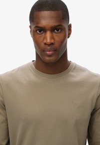 Koszula z długim rękawem w jasnym oliwkowym kolorze z bawełny, z okrągłym dekoltem i małym haftowanym logo na lewej piersi. Gładka tekstura.