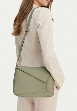 Donna con tailleur beige chiaro porta una borsa a tracolla in pelle verde oliva con tracolla regolabile sulla spalla.