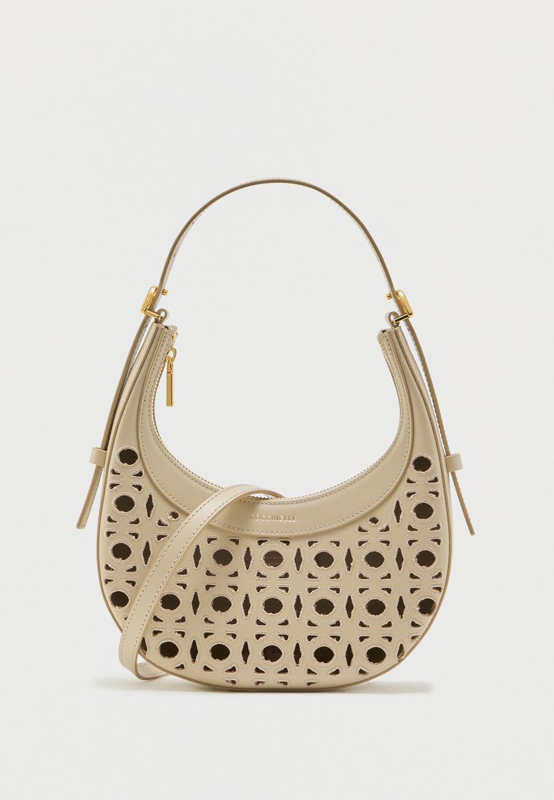 Μπεζ δερμάτινη τσάντα σε σχήμα ημισελήνου με διάτρητα γεωμετρικά μοτίβα, λαβή στο πάνω μέρος και ρυθμιζόμενο ιμάντα ώμου σε λευκό φόντο.
