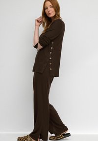 Conjunto de loungewear de punto acanalado marrón que incluye una camiseta de manga larga con botones laterales y pantalones de pierna ancha; zapatillas de leopardo complementan el conjunto.