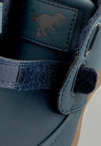 Le détail de la chaussure en cuir bleu présente une sangle velcro, un accent souple texturé et un petit logo de dinosaure en relief sur le côté.