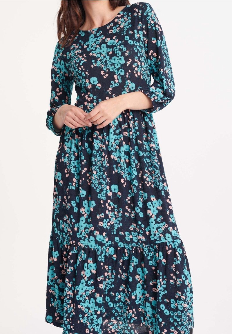 Femme portant une robe bleu marine à manches longues, coupe ample, avec un motif floral turquoise et rose clair.