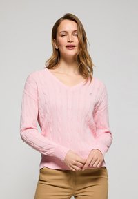 Ljusrosa V-ringad stickad tröja med kabelstickat mönster, ribbade ärmslut och nederkant. Modellen poserar med händerna på höfterna, iförd beige byxor.