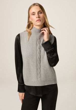 Grauer gestrickter Sweater-Weste mit hohem Kragen und einem Zopfmuster in der Mitte, getragen über einem schwarzen Hemd mit Kragen.