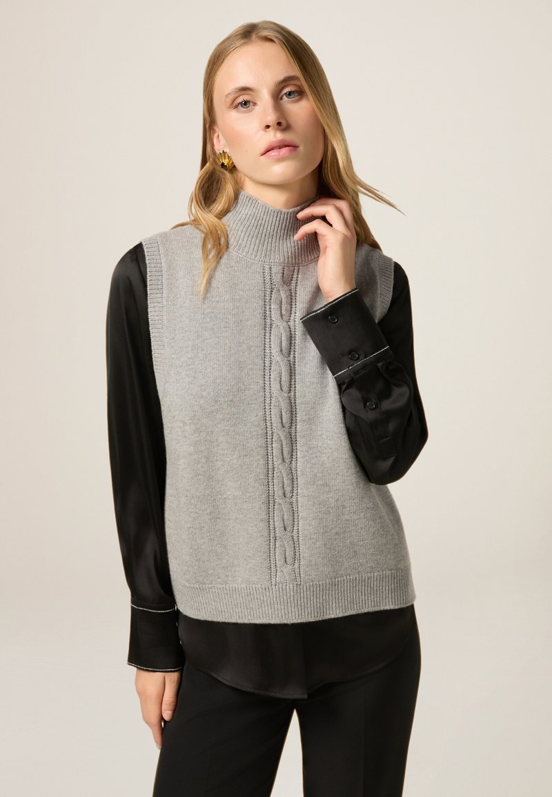 Chaleco de punto gris con un cuello alto y un patrón de punto trenzado en el centro, llevado sobre una camisa negra con cuello.