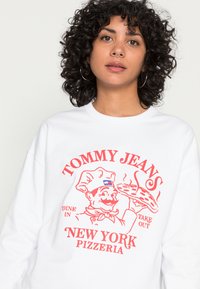 Biela mikina s červenou grafikou kuchára držiaceho pizzu, text znie "TOMMY JEANS NEW YORK PIZZERIA" v tučnom písme.