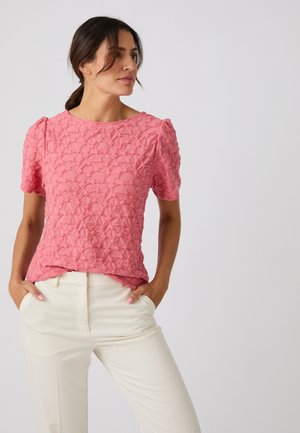 Frau mit dunklem Haar in einer rosa strukturierten Kurzarmbluse und weißen Hosen, die mit den Händen in den Taschen zur Seite schaut.