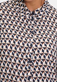 Košilové tričko s geometrickým vzorem v béžové, námořnické modré a hnědé barvě. Vyrobeno z hladké tkaniny s klasickým límcem a zapínáním na knoflíky.