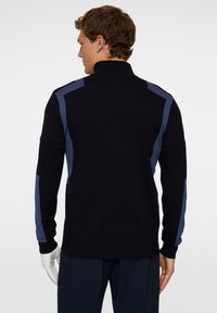 Svart tröja med hög krage, med blå accentpaneler på axlarna och ärmarna. Stickad struktur med ribbade muddar och nederkant.