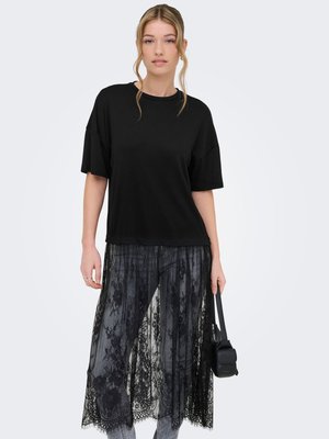 Młoda kobieta o blond włosach, ubrana w czarny oversize t-shirt i czarną koronkową spódnicę midi, trzymająca małą czarną torebkę.