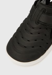 Juoda sandalė su tekstūruotu viršumi su bangų raštais, reguliuojama diržele, ventiliacijai skirtomis skylutėmis ir balta pada.