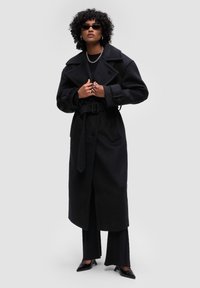 Černý vlněný trenčkot s over-sized klopami, páskem v oblasti pasu a ladícími knoflíky. Stylizováno s upnutými černými kalhotami a lodičkami s špičatou špičkou.