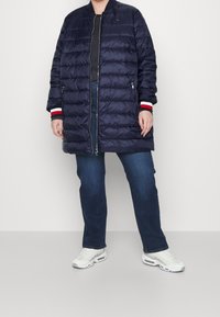 Námořnický péřový kabát s horizontálním prošíváním, žebrovanými manžetami s červenými a bílými detaily, zapínáním na zip a bočními kapsami, nošený přes tmavé džíny.