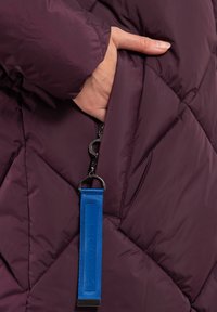 Bordeaux pufferjack met een diagonaal gestikt patroon, zijzak met rits en een blauwe merklabel aan het stof bevestigd.