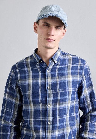 Μπλε καρό πουκάμισο με λευκές και ανοιχτό γκρι λεπτομέρειες, συνδυασμένο με ένα ανοιχτό μπλε τζην καπέλο με φθαρμένες άκρες.