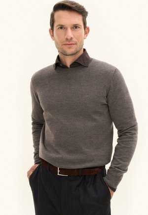Mann mit kurzen braunen Haaren, der einen grauen Pullover über einem dunklen Hemd, schwarze Hosen und einen braunen Gürtel trägt, steht mit den Händen in den Taschen.