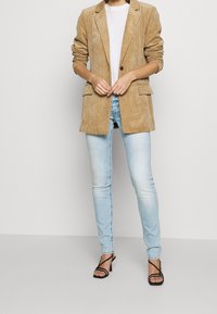 Beige Corduroy-Blazer mit zwei Fronttaschen, kombiniert mit hellblauen Skinny-Jeans. Ausgestattet mit schwarzen Riemchen-Heels. Minimalistischer Stil.