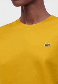 Personne portant un t-shirt jaune à col rond avec un petit logo de crocodile vert sur la poitrine gauche.