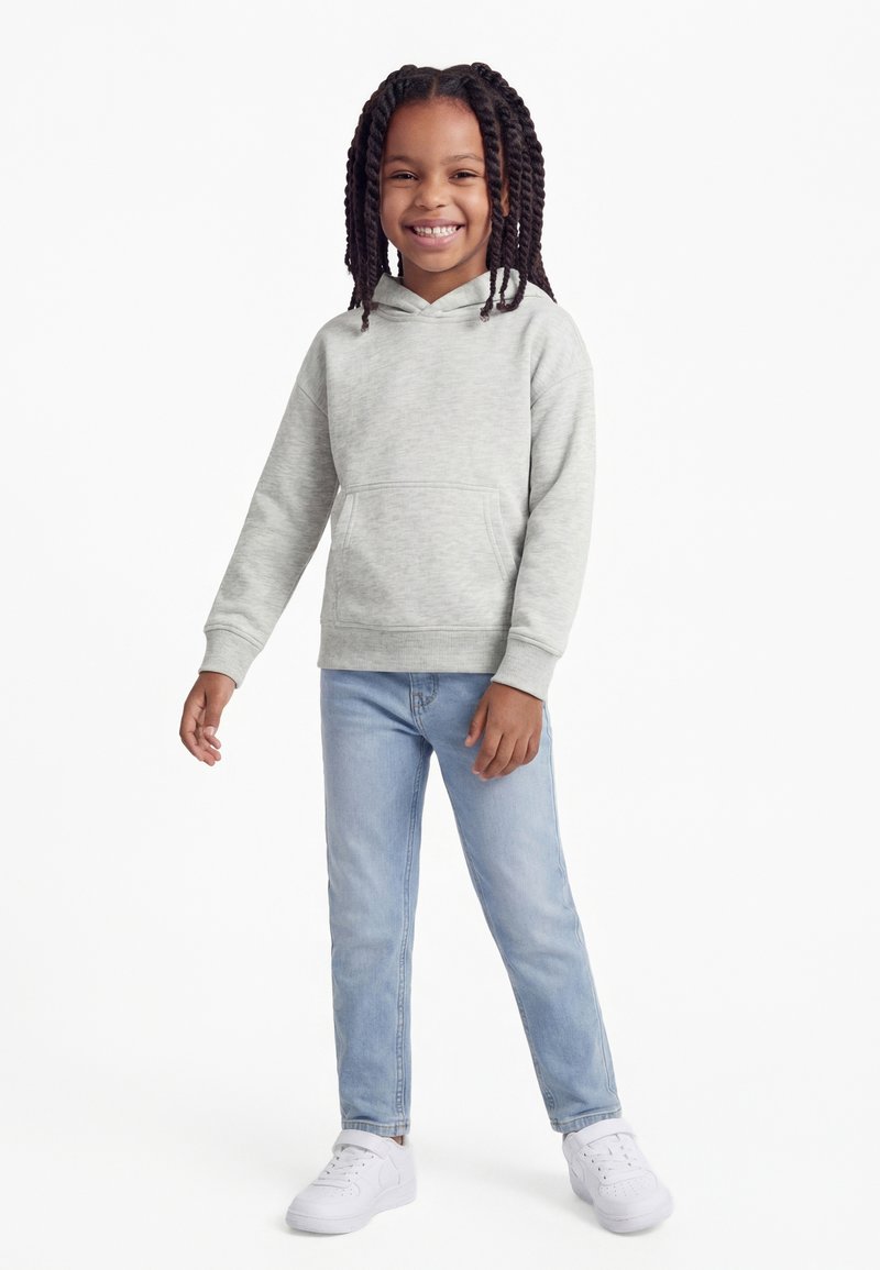 Bambino con capelli intrecciati che sorride, indossa una felpa con cappuccio grigio chiaro, jeans azzurri e sneakers bianche, in piedi davanti a uno sfondo bianco.