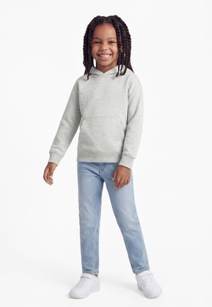 Enfant avec des cheveux tressés souriant, portant un sweat à capuche gris clair, un jean bleu clair et des baskets blanches, debout devant un fond blanc.