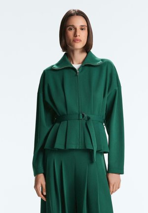 Donna con capelli castani lisci, indossa una giacca verde scuro con zip e cintura abbinata a una gonna a pieghe, in piedi su uno sfondo chiaro uniforme.