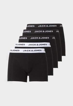 Schlanker dunkelhäutiger Mann steht barfuß in schwarzen Jack & Jones Boxershorts vor einfachem hellem Hintergrund und schaut zur Seite.
