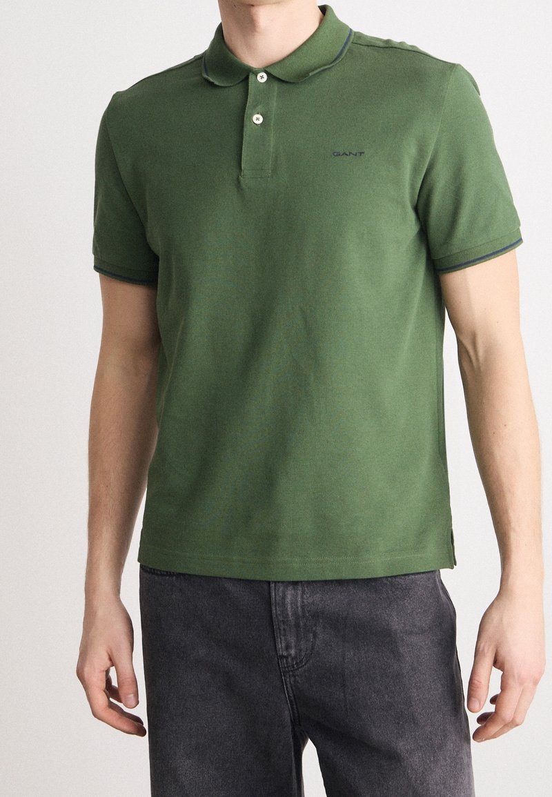 Homme portant un polo vert à manches courtes avec des boutons et un pantalon gris foncé, debout les bras détendus.